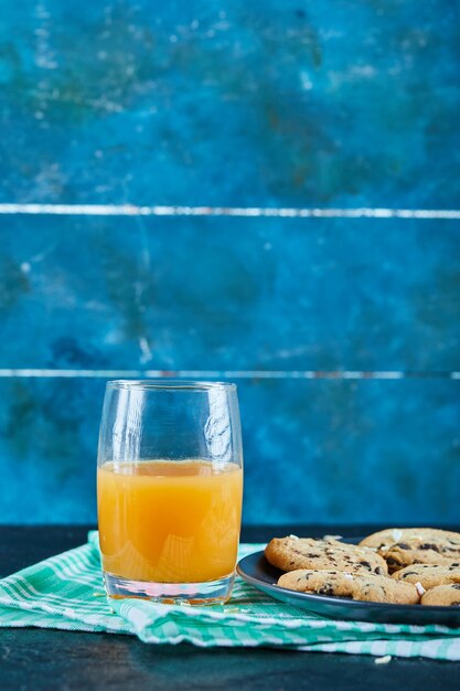 暗いテーブルの上のチョコレートチップクッキーのプレートとオレンジジュースのガラス
