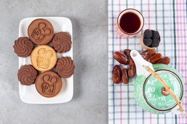 Plate of cookies next to ornate teapot, dates, a cup of tea and a bowl of tea leaves on marble background. High quality photo