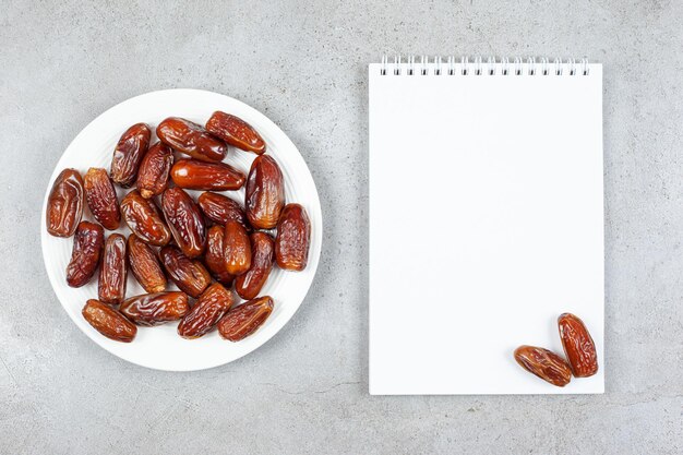A plate of fresh dates next to a notebook with two dates on it on marble background. 