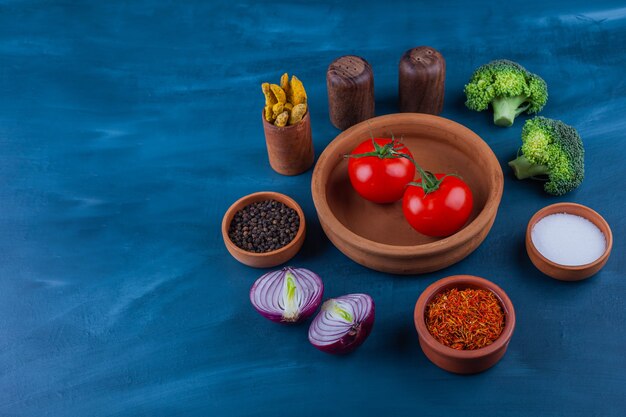 Plate of fresh tomatoes, onion, broccoli and condiments on blue surface.