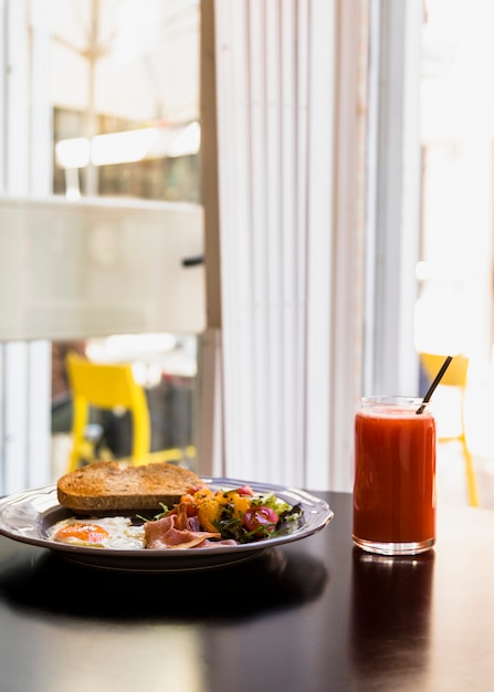 Бесплатное фото Тарелка тостов; салат; жареные яйца; бекон со стаканом сока на черном столе возле окна