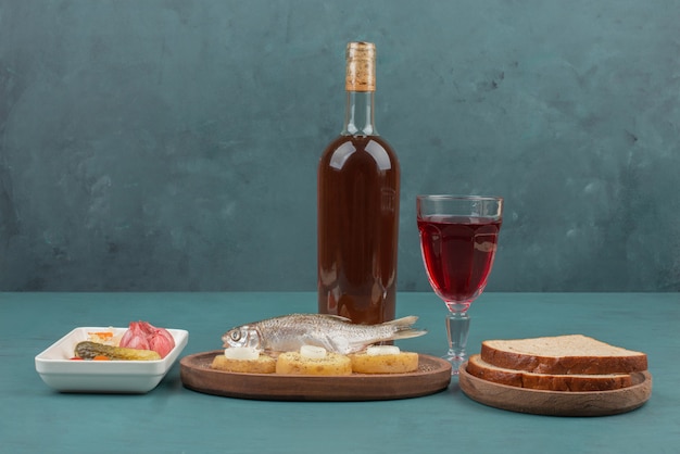 Plate of pickled vegetables, fish, slices of bread and red wine on blue table.