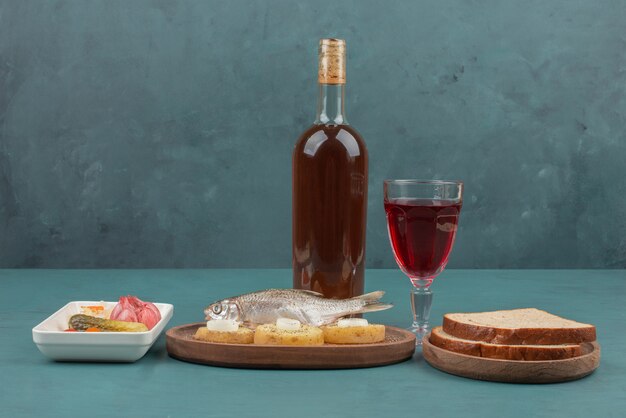 Plate of pickled vegetables, fish, slices of bread and red wine on blue table. 