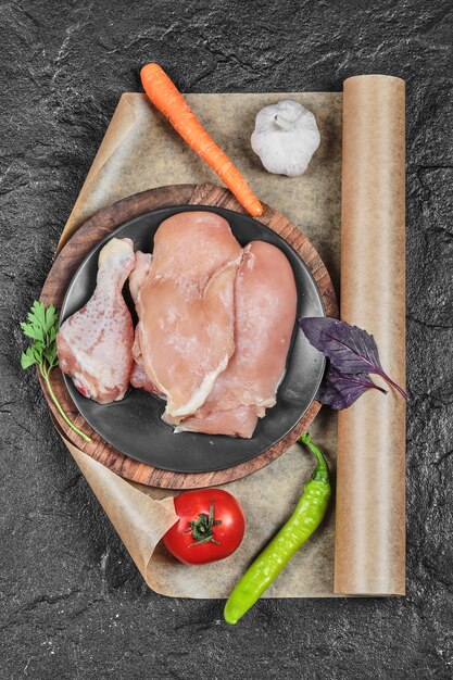 Plate of raw chicken parts with tomato and carrot on dark surface