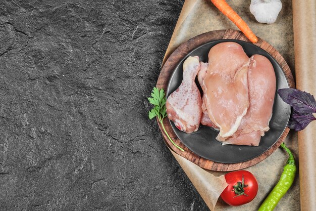 Plate of raw chicken parts with tomato and carrot on dark surface