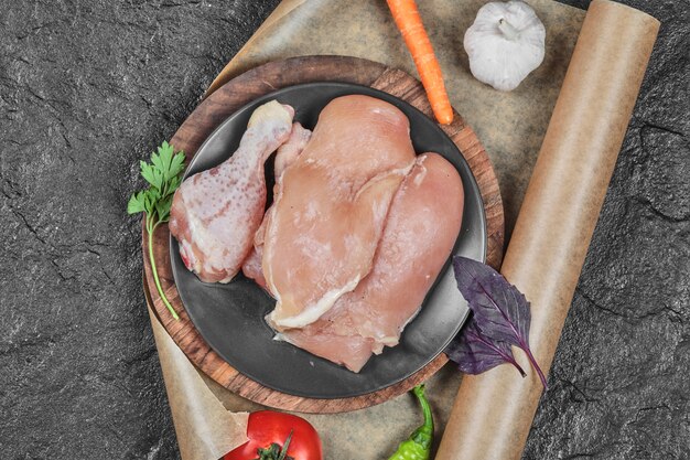 Plate of raw chicken parts with tomato and carrot on dark surface