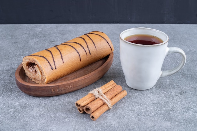 Plate of roll cake, cup of tea and cinnamon sticks on marble surface
