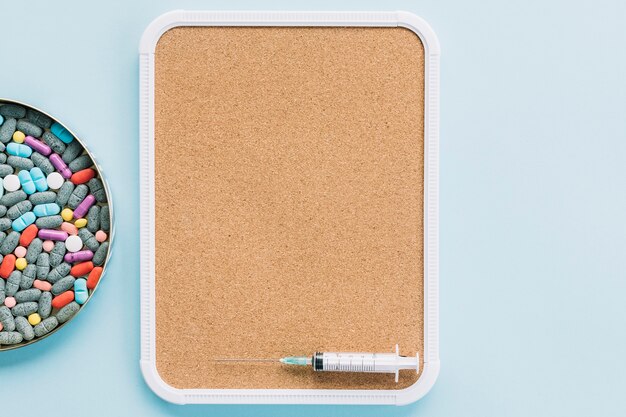 Plate with colorful pills and syringe in cork tray against blue backdrop