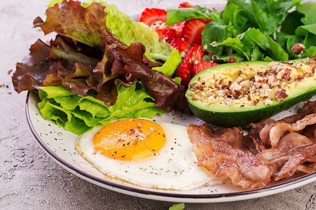 Тарелка с кето диетическое питание. Жареные яйца, бекон, авокадо, руккола и клубника. Кето завтрак.