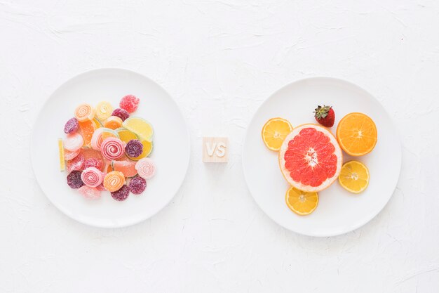 Plates of sweet candies versus fruits over white texture rough background