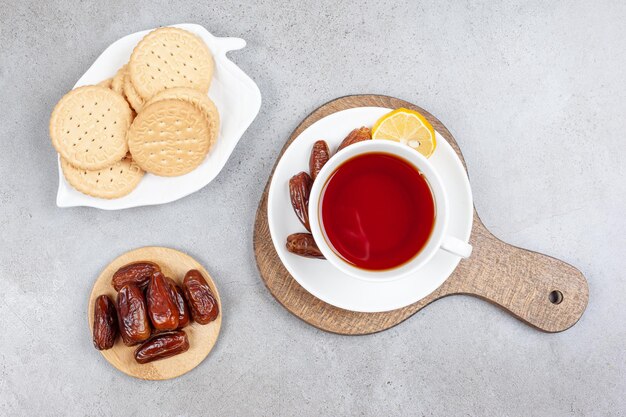 A platter of cookies and a small pile of dates next to a cup of tea with some dates on saucer on a wooden board, on marble surface