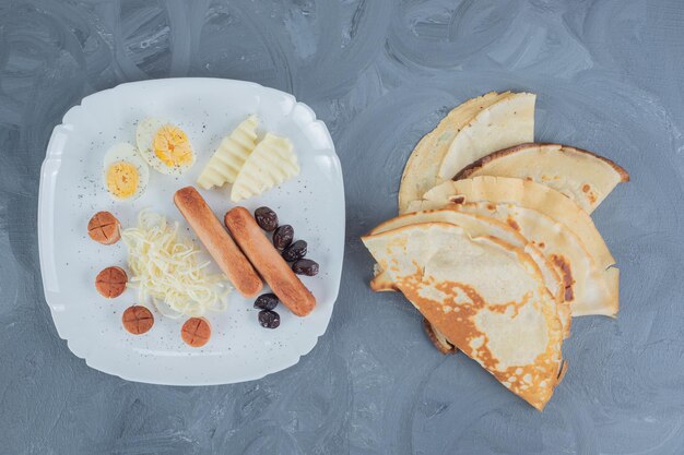 Platter of eggs, cheese, olives and sausages next to pancakes on marble table.