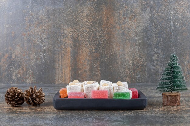 Platter of marmelades and turkish delights next to a pine cone and a tree figurine on wooden surface