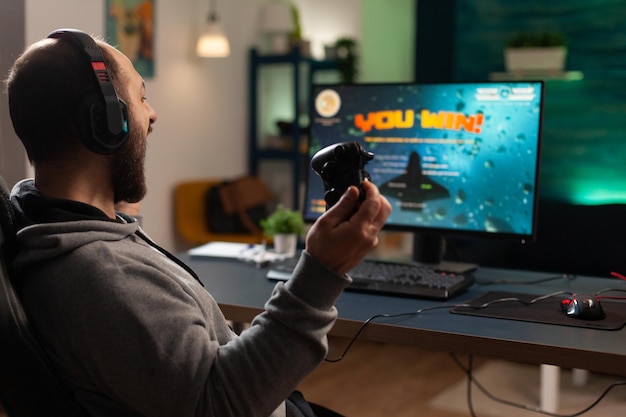 Player winning video games with controller and headset in front
of monitor. man using joystick and headphones, playing online games
on computer. person celebrating game win for leisure.