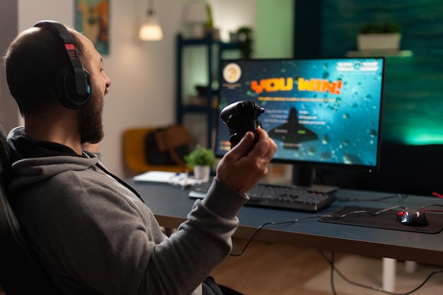 Player winning video games with controller and headset in front of monitor. Man using joystick and headphones, playing online games on computer. Person celebrating game win for leisure.