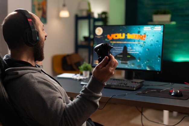 플레이어가 모니터 앞에 컨트롤러와 헤드셋을 사용하여 비디오 게임을 이기고 있습니다. 조이스틱과 헤드폰을 사용하여 컴퓨터에서 온라인 게임을 하는 남자. 여가를 위해 게임 승리를 축하하는 사람.