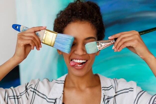 Playful African  female artist holding brushes and making grimaces, showing tongue.