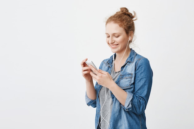 Playful attractive redhead female with bun, laughing while looking at screen of smartphone, texting and listening music in headphones