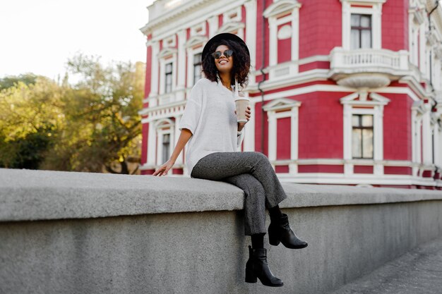 Playful black woman with Afro hairs sitting on the bridge and having fun. Wearing leather boots and whoop  trendy trousers . Travel mood. Happy leisure time in old European city.