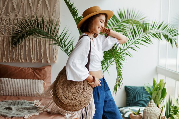 Playful bohemian woman posing in stylish bedroom with amazing interior, palms and macrame