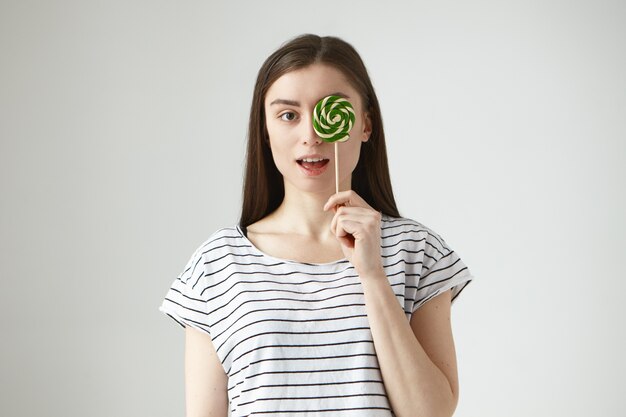 Playful brunette girl wearing stylish striped t-shirt having fun indoors, opening mouth widely and covering one eye with colorful round hard candy. Pretty young woman enjoying lollipop 