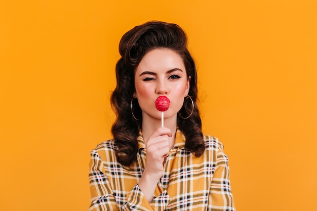 Playful brunette lady licking lollipop. Studio shot of amazing pinup girl in checkered shirt eating candy.