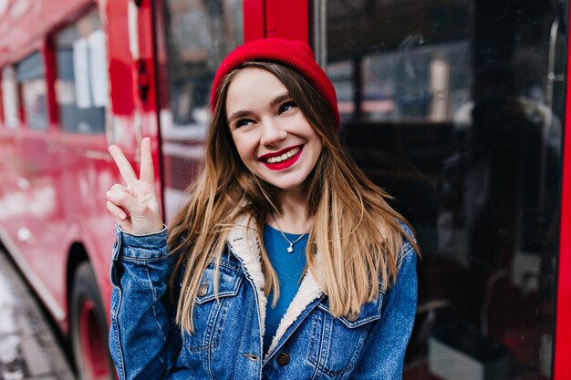 Playful caucasian girl with trendy hairstyle posing with peace sign. Outdoor shot of pretty woman in denim jacket laughing during photoshoot.