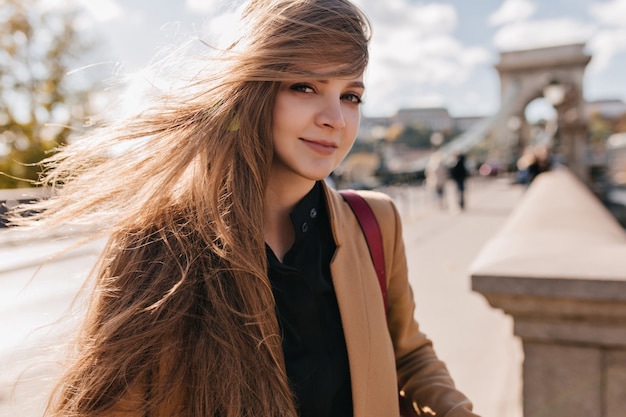 Playful fair-haired woman in beige jacket walking outdoor in windy day