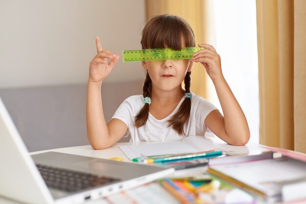 Playful female child in white t shirt sitting at table in front of opened notebook, covering eyes with green ruler, distance education during quarantine, pointing finger up, having idea.