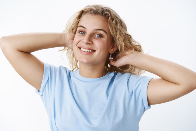 Playful, feminine attractive european woman with short blond curly hairstyle holding hands behind head as combing hair smiling joyfully and gazing at camera carefree and relaxed over white wall