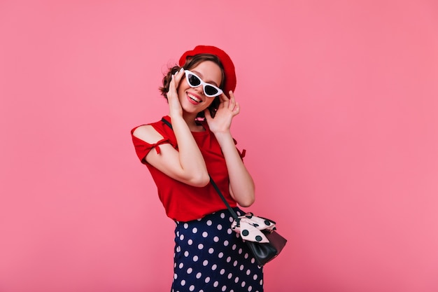Foto gratuita donna francese allegra che posa in occhiali da sole. attraente ragazza dai capelli scuri in berretto rosso sorridente sul muro roseo.