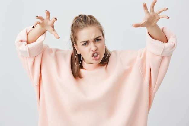 Playful funny girl dressed in pink sweatshirt, frowning her face and showing her teeth, holding her arms like she is trying to scary someone, posing . Fun, people, lifestyle.