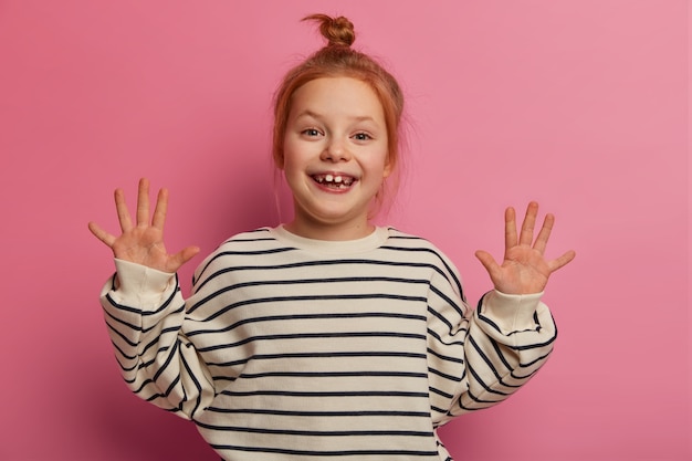 Playful funny red head girl raises both palms , smiles pleasantly, has missing teeth, wears striped jumper, poses against rosy pastel wall, has carefree expression. Childhood concept