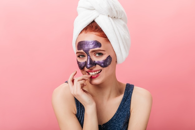 Playful girl with towel on head looking at camera on pink background. Studio shot of charming caucasian woman with face mask.