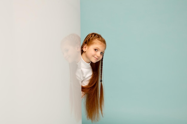 Playful. happy kid, girl isolated on blue studio background. looks happy, cheerful. copyspace for ad. childhood, education, emotions, facial expression concept. peeking out from behind the wall.
