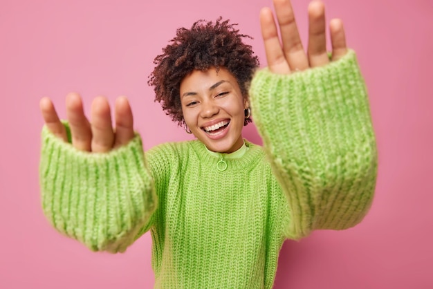 Playful happy woman keeps hands forwards camera smiles joyfully wears warm knitted jumper expresses positive emotions amuses someone isolated over pink background. Cheerful curly girl feels glad