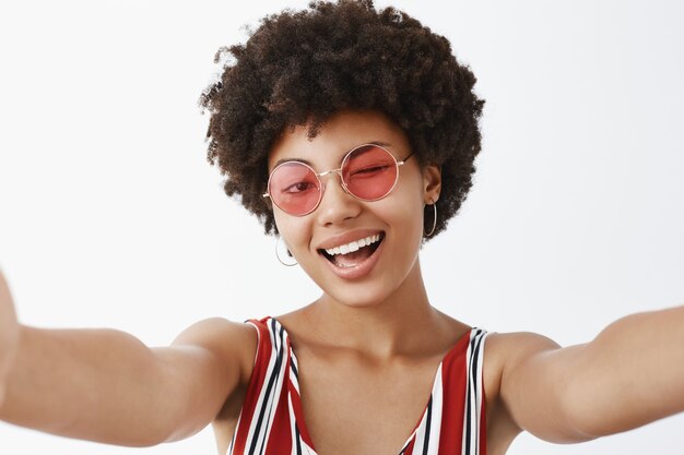 Playful hot African American with afro hairstyle, pulling hands towards to make selfie, winking joyfully and smiling broadly, making new profile pic for social network