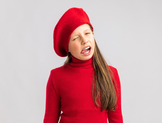 Playful little blonde girl wearing red beret looking at camera showing tongue winking isolated on white wall with copy space