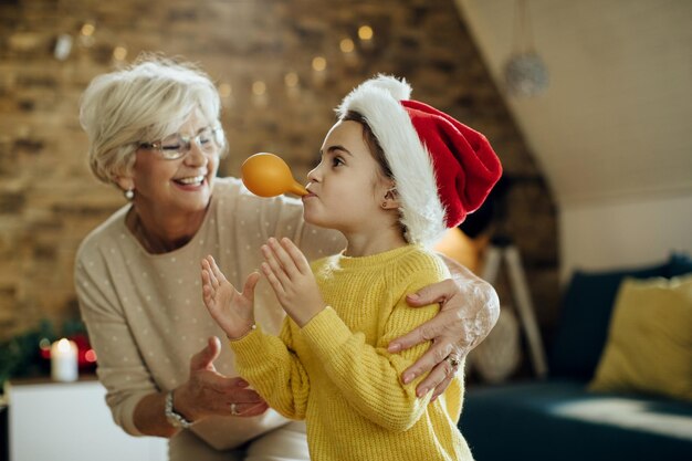 Playful little girl and her grandmother blowing balloon and having fun on Christmas day