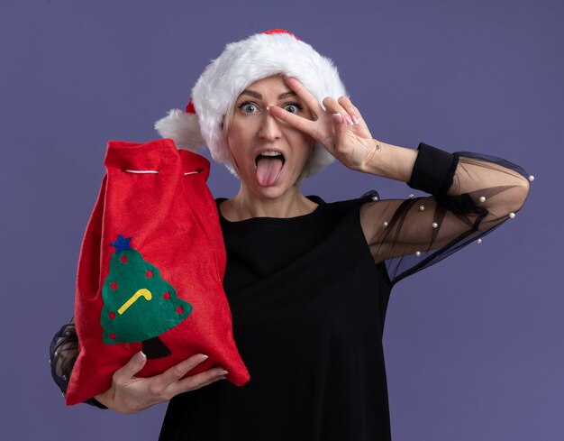 Игривая блондинка средних лет в рождественской шляпе с рождественским мешком смотрит в камеру, показывая язык и символ v-знака возле глаза, изолированную на фиолетовом фоне