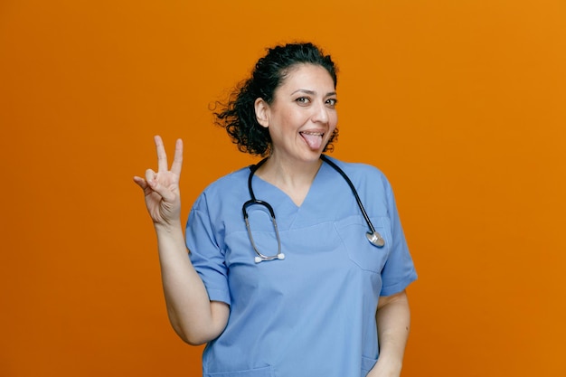 Игривая женщина-врач средних лет в униформе и со стетоскопом на шее смотрит в камеру, показывающую знак мира и язык, изолированный на оранжевом фоне