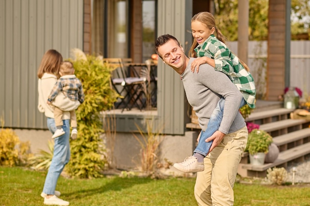 Playful mood. Smiling young adult man in casual clothes holding enthusiastic school-age daughter on back and woman with child with back to camera in courtyard of country house