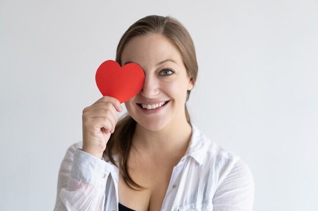 Playful pretty woman holding paper heart in front of her eye