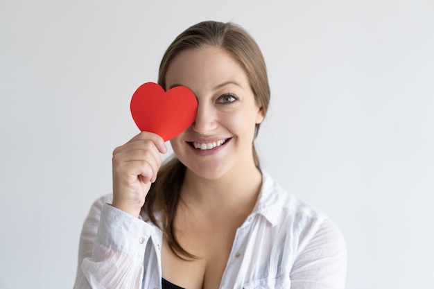 Playful pretty woman holding paper heart in front of her eye