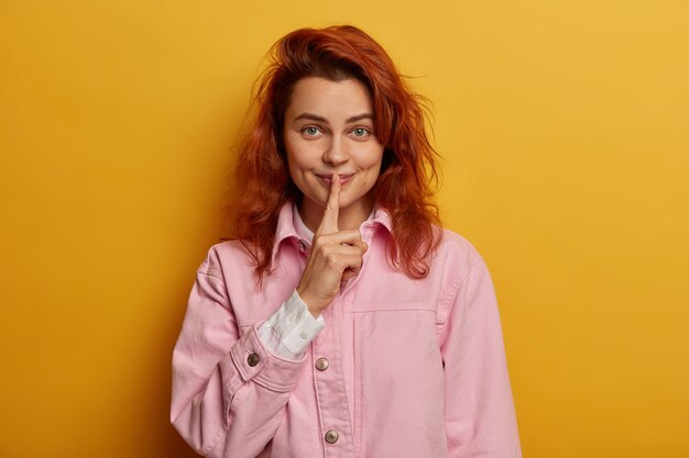 Playful redhead pretty woman with satisfied secret look, makes hush gesture, gossips with friend, dressed in rosy denim jacket