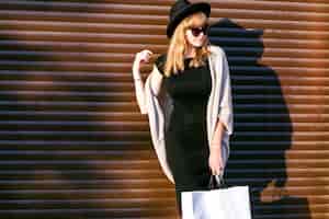 Foto gratuita giocosa donna timida che nasconde il pacchetto della spesa, ridendo timido. carina donna sorridente felice attraverso le mani. cappello nero, cappotto grigio, vestito nero, vestiti autunnali. caldo stile invernale