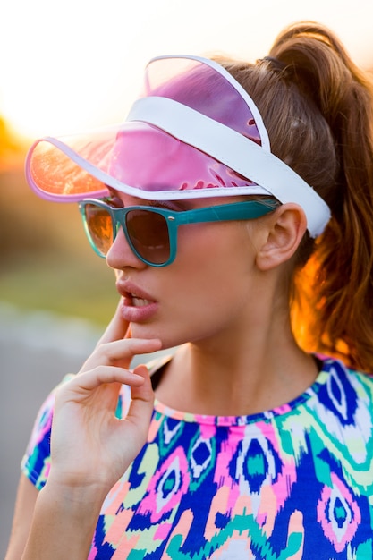 Playful slim girl in stylish colorful crop top, transparent cap and sunglasses posing on the street at sunset.