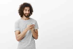 Free photo playful thoughtful handsome guy with curly hair, holding smartphone, smirking and looking with curious intrigued expression