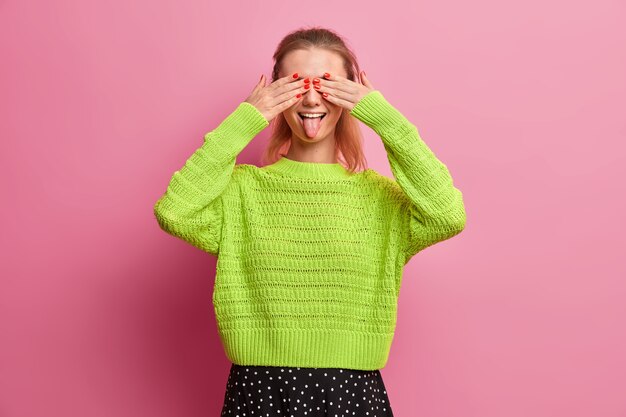 Playful woman covers eyes and strikes out tongue, has fun and foolishes around, plays with her younger sister, dressed in loose knitted green jumper