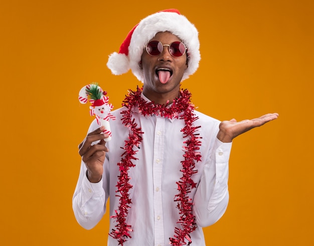 오렌지 벽에 고립 된 혀와 빈 손을 보여주는 사탕 지팡이 장식을 들고 목 주위에 반짝이 갈 랜드와 산타 모자와 안경을 착용 장난 젊은 아프리카 계 미국인 남자